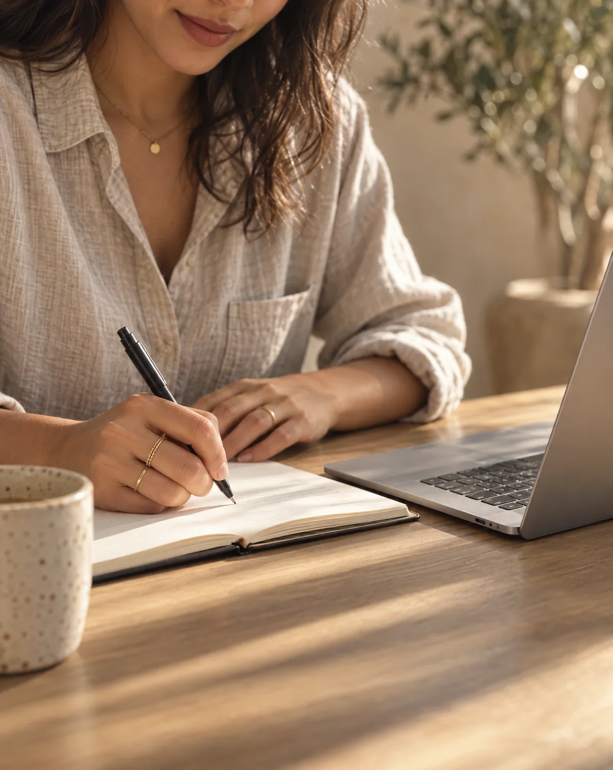 Close-up of a woman writing notes in a notebook beside an open laptop on a sunlit oak table.